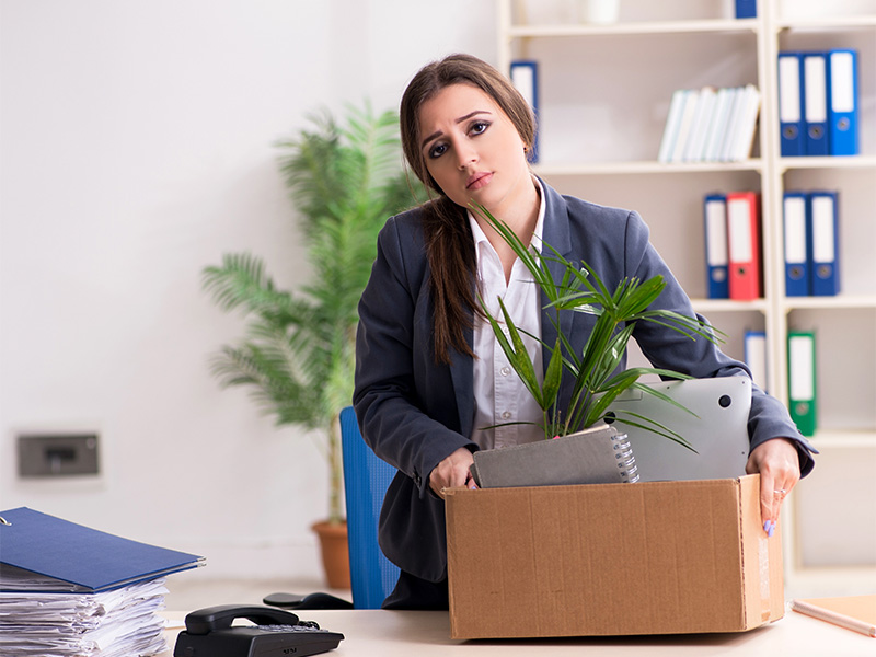 Eine Angestellte verlässt nach Kündigung das Büro mit Karton voller persönlicher Gegenstände – Symbol für Kündigung