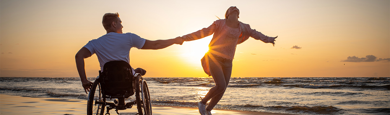 Mann im Rollstuhl am Strand mit Partnerin, zeigt Mobilitätshilfen und Hilfsmittel für Alltag und Pflege.